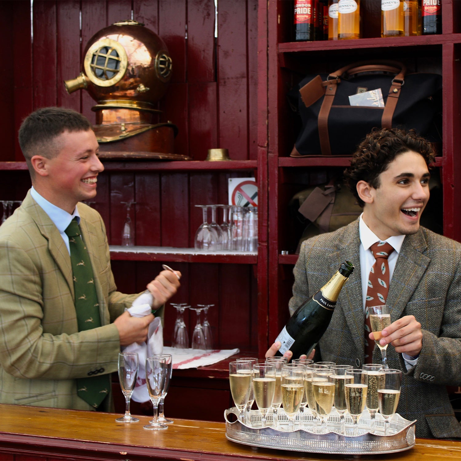 Two men pouring champagne at the Dubarry stand at the Game Fair in 2023. Both men are wearing grey and green tweed jackets; one is wearing a green tie, while the other wears a red tie.//© Dubarry of Ireland