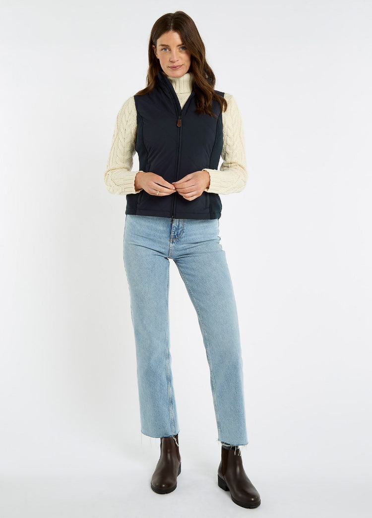 Woman wearing a navy quilted vest over a cream cable knit sweater with jeans and brown leather ankle boots