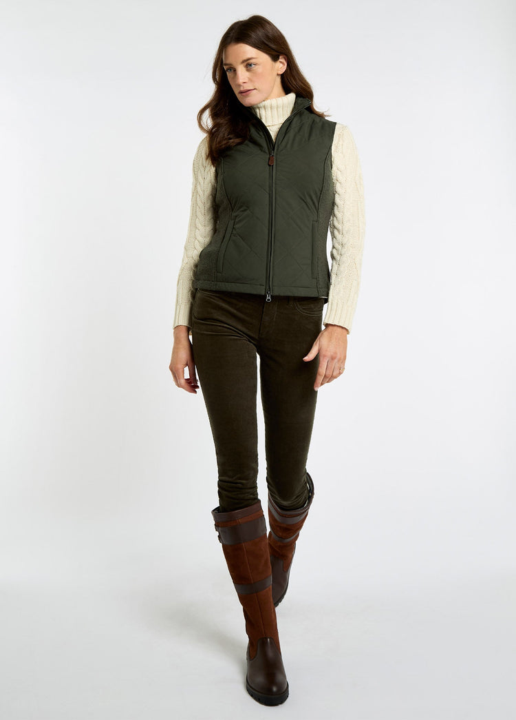Woman wearing an olive quilted vest over a cream cable knit sweater with brown trousers and tall leather country boots