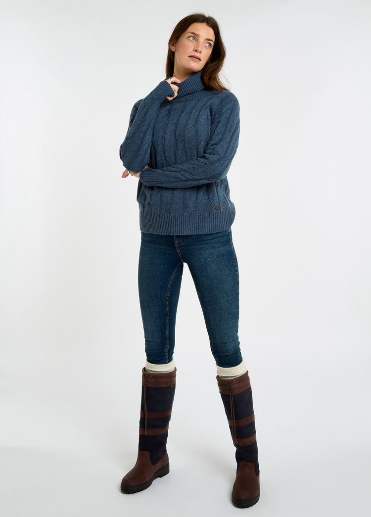 Woman wearing an indigo roll neck knit sweater with jeans and tall brown country boots in a studio setting