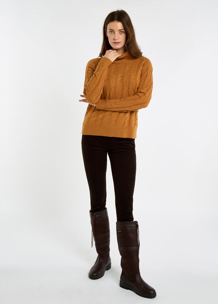 Woman wearing an ochre roll neck cable knit sweater with dark trousers and tall brown boots, styled for country wear