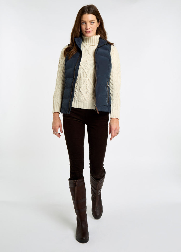 Woman wearing a navy quilted vest over a cream cable knit sweater with brown trousers and tall leather country boots