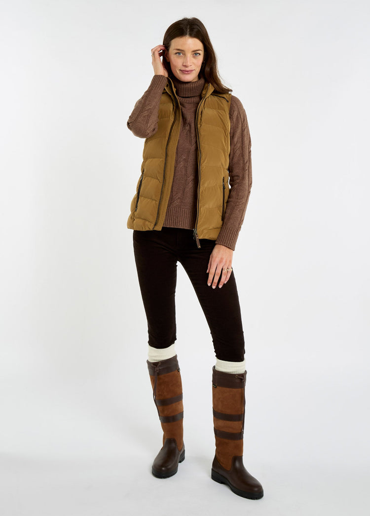 Woman wearing an ochre quilted vest over a brown sweater, black trousers and tall brown country boots.