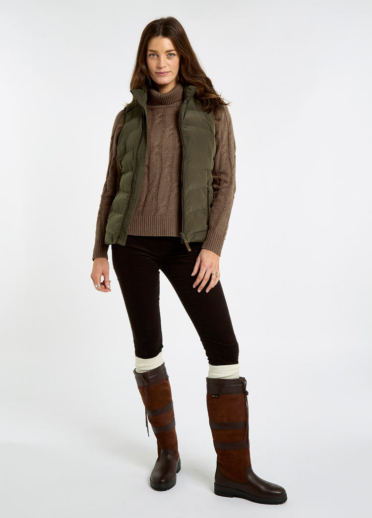 Woman wearing an olive quilted vest over a brown knit sweater with dark trousers and tall brown country boots in a studio setting