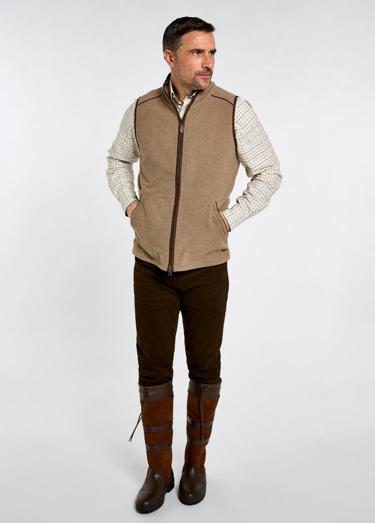 Man wearing an elk brown fleece vest over a checked shirt with dark trousers and tall leather country boots pose.