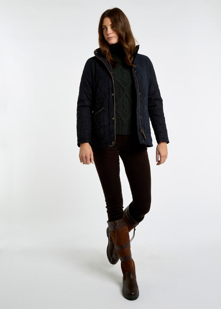 Woman wearing a navy quilted jacket with herringbone texture, layered over a dark cable knit sweater and paired with brown riding boots