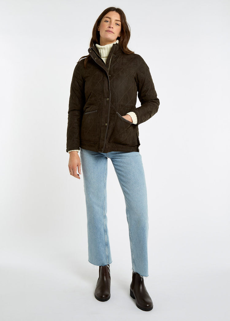 Woman wearing an olive herringbone quilted jacket with a high collar and pockets styled with jeans and brown ankle boots