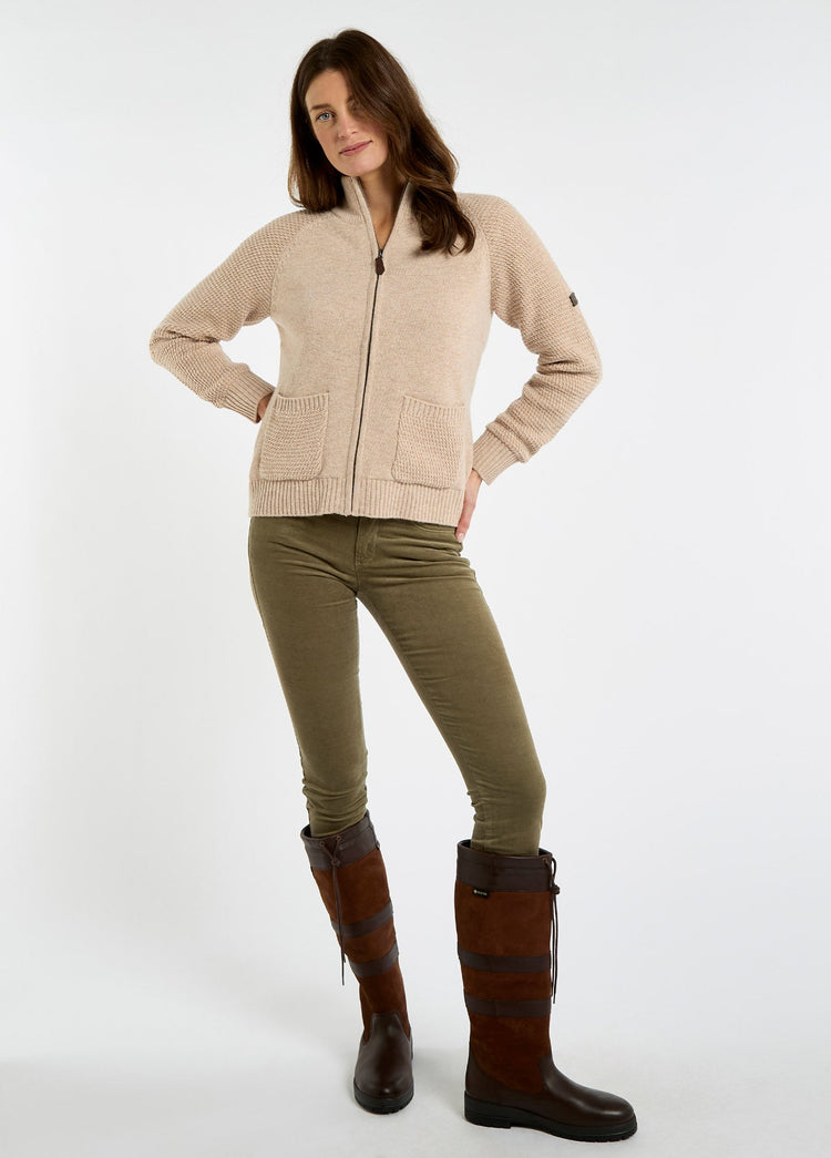 Woman wearing an oat full zip knit sweater with front pockets olive trousers and tall brown leather country boots