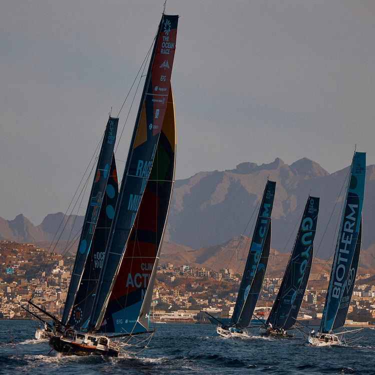 Square image of five ships sailing at sea, with the town of Mindelo in the background, as they set off on the Ocean Race second leg 2023.//© GUYOT environment - Team Europe | Charles Drapeau