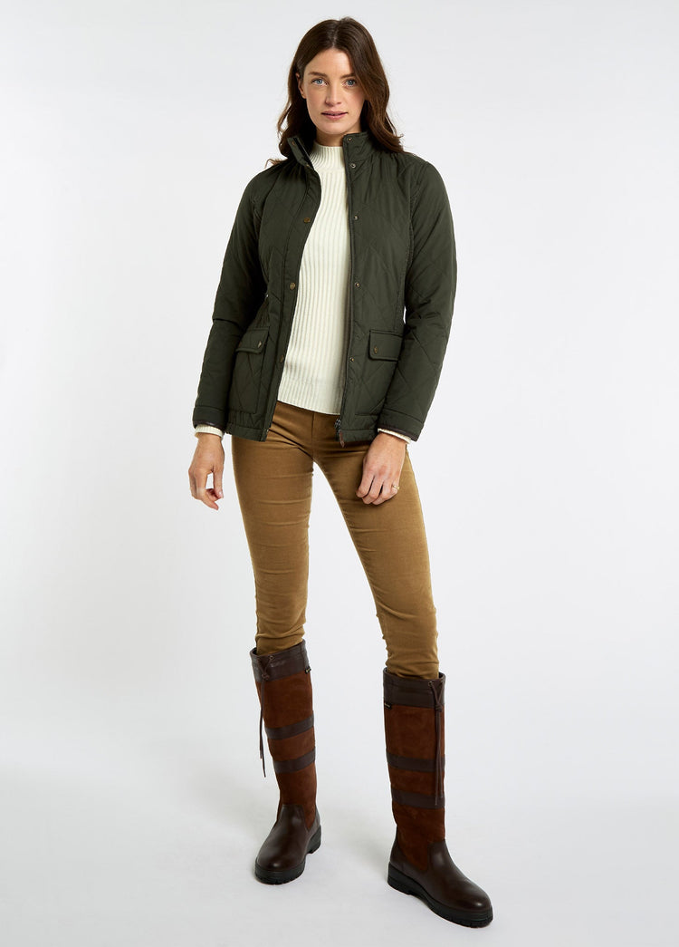 Woman wearing an olive lightweight jacket with knit side panels, cream sweater, brown trousers and tall leather boots against a white background