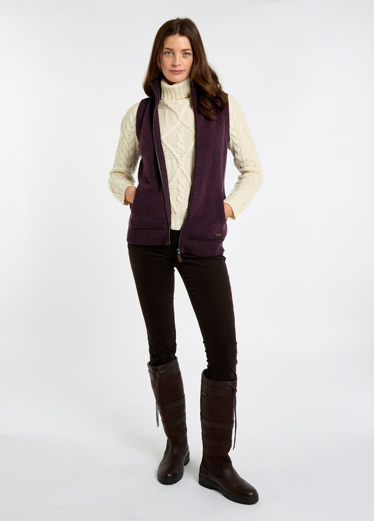 Woman wearing a damson sleeveless full zip knitwear vest over a cream cable knit sweater with dark trousers and tall brown boots