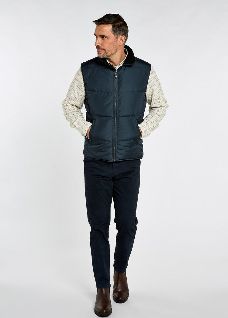 Man wearing a navy quilted vest over a cream patterned sweater, with dark trousers and brown leather boots