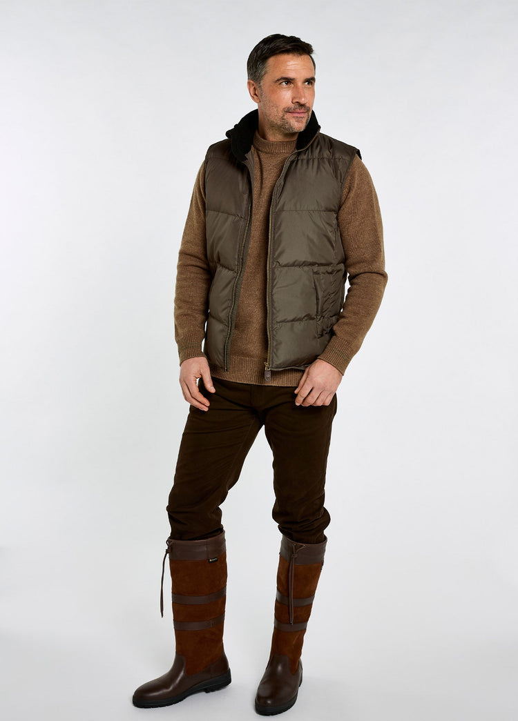 Man wearing an olive quilted vest over a brown knit sweater with dark trousers and tall brown country boots
